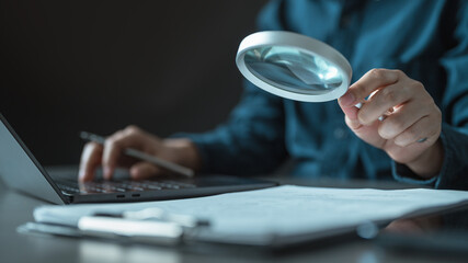 Fototapeta premium audit, documents, magnifying, glass, verification, Close-up of a person using a magnifying glass to inspect documents beside a laptop, representing auditing, verification, and detailed analysis.