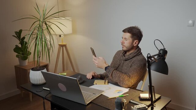 Man sitting at desk using smartphone while working in a home office with laptop and documents. Tired freelancer procrastinating at workplace during remote work. Unproductive man using phone