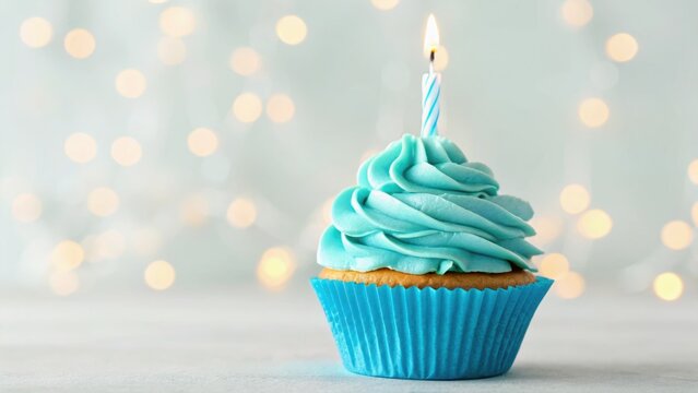 A decorative cupcake with blue frosting and a lit candle, set against a soft, blurred background of warm lights.