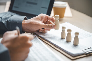 Close-up of hands evaluating wooden human figures on documents, representing hiring decisions,...