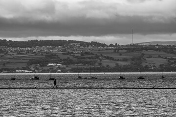 Person walking beside boats on coastal water in black and white
