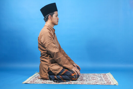 side view of a Muslim man sitting in prayer position on a prayer mat isolated blue background