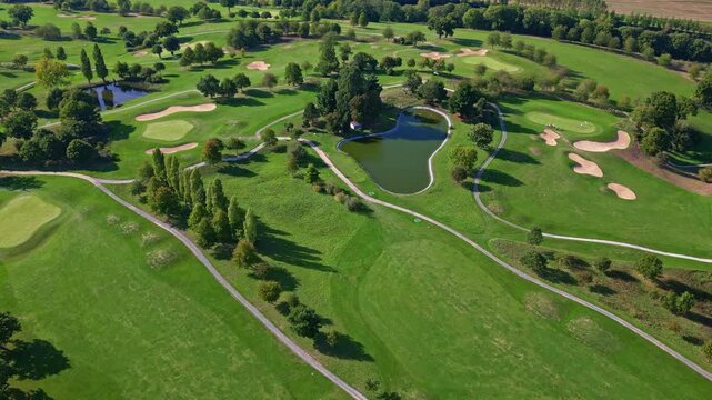 Drone glides slowly above a sprawling summer golf course, revealing smooth greens, rolling hills, sculpted sand traps, and reflective ponds framed by leafy deciduous and evergreen trees
