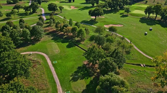 Drone glides over golf course featuring tree-lined fairways with a mix of tree species&mdash;including oak, maple, and ash alongside patches of evergreen pine and ornamental willow near water features.