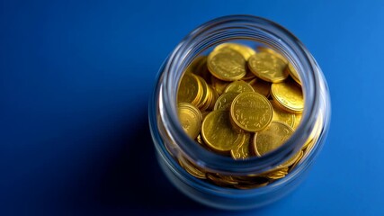 A glass jar filled with shiny gold coins is shown on a blue background. The focus is on the importance of saving money and budgeting wisely. Visuals highlight the jar's contents