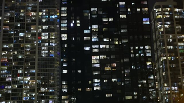Close up nighttime view of tall residential high-rise buildings with many illuminated windows. Each window reveals a glimpse of life inside, creating a vibrant urban mosaic.