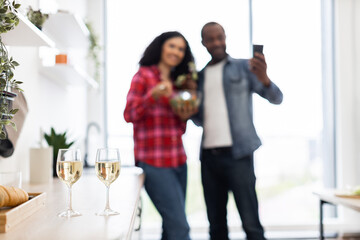 A couple is in their kitchen, with the man taking a selfie while the woman holds a bowl. Two glasses of wine are on the counter.