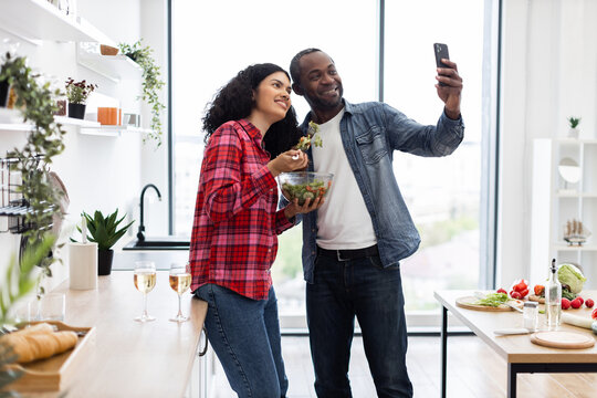 A diverse couple smiles while taking a selfie in their modern kitchen, enjoying a salad and wine.