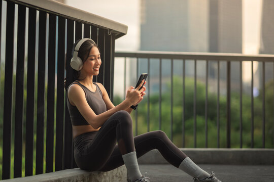 Asian woman relaxing after workout with wireless headphones and smartphone, active lifestyle in urban city, fitness break, listening to music, checking phone, healthy routine, outdoor exercise morning