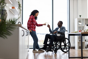 A couple is toasting with wine glasses in a modern kitchen. One person is in a wheelchair.