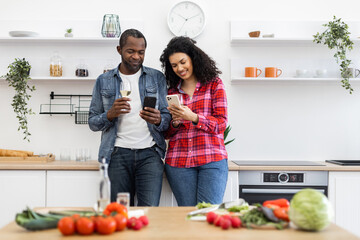 A diverse couple smiles while looking at their phones in a modern kitchen, with food on the counter.