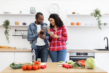 A diverse couple enjoys wine and uses their phones in a modern kitchen setting, surrounded by fresh produce.