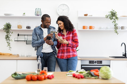 A diverse couple enjoys wine and uses their phones in a modern kitchen setting, surrounded by fresh produce. - Powered by Adobe