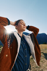 Naklejka premium Woman, portrait and outdoors with sunlight and jacket near mountains, enjoying nature and travel. Female model in denim shirt with closed eyes, relaxed pose in countryside golden hour.