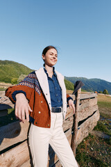 Naklejka premium woman countryside fence mountains jacket smiling outdoors portrait. Young woman stands leaning on a wooden fence in a rural landscape wearing a patterned jacket, smiling confidently under clear blue
