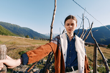 Naklejka premium Woman portrait in mountains countryside by wooden fence wearing warm jacket and denim shirt, standing outdoors with fashion look and scenic valley landscape under clear sky.