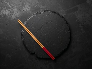 Wooden chopsticks with red tips placed diagonally on round black slate plate.