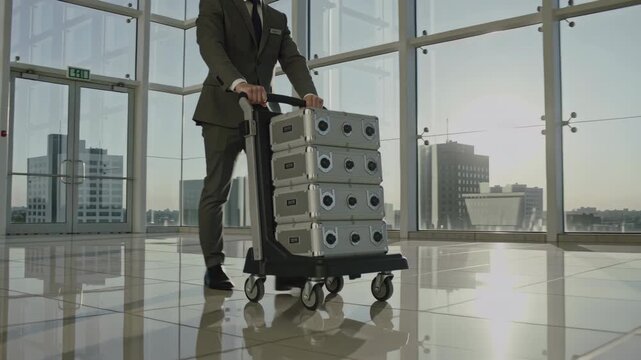Businessman in formal suit pushing trolley with stacked metal cases through modern glass office lobby during sunrise, reflecting sunlight on polished floor, showcasing urban cityscape