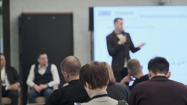Audience watches blurred presenter in formal attire delivering talk at corporate training event with projector screen