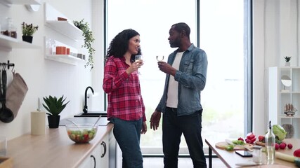 A diverse couple enjoys a glass of wine while chatting in a modern kitchen setting, with a salad bowl on the counter. - Powered by Adobe