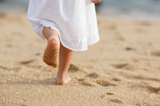 Child with sandy bare feet walking on the beach in a white dress