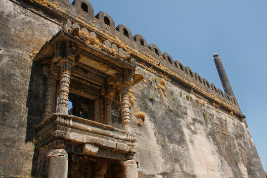 Ancient stone wall and carved balcony structure at Uparkot Fort in Junagadh, Gujarat. Historic fort architecture with weathered textures under a clear blue sky.