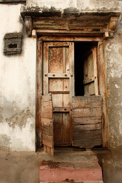 Old rustic wooden door with broken lower panels on a weathered wall in Saurashtra, India. Aged textures and earthy tones reflect rural heritage and traditional village architecture.