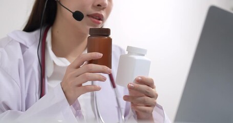 Asian female doctor wearing headset holding medication bottles while consulting online with patient, explaining drug usage through telehealth platform, modern remote healthcare technology concept