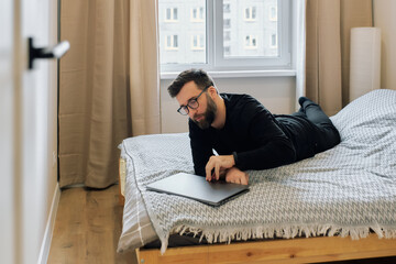 Man working on laptop while relaxing on bed in cozy bedroom during daytime