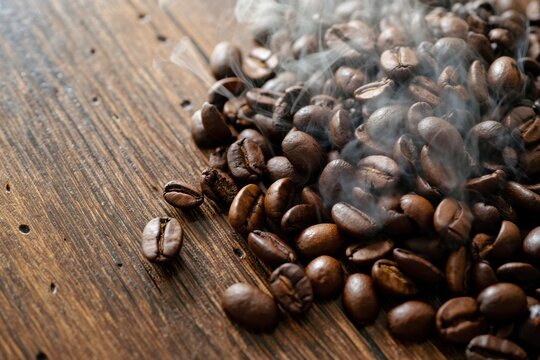Coffee beans scattered on a wooden surface with steam curling upward, aromatic.