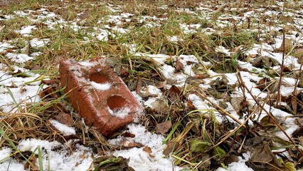 Red brick partially covered in snow and surrounded by fallen leaves and grass, showcasing the contrast of nature and man-made materials in a winter landscape