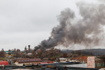 A large fire burns in an industrial or semi-urban area, sending thick, dark smoke high into the cloudy sky. Flames can be seen among trees and buildings.