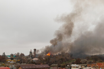 A large fire burns in an industrial or semi-urban area, sending thick, dark smoke high into the cloudy sky. Flames can be seen among trees and buildings.