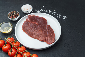 Raw beef steak on white plate with cherry tomatoes, pepper, salt and oil on black background