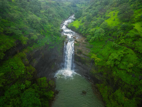 Aerial view of a stunning waterfall in Bhandardara surrounded by lush green hills, flowing into a serene pool during the monsoon season.