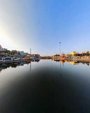 A wide panoramic view of Nashik&rsquo;s lakeside ghats with clear blue sky, colorful buildings, and mirror-like reflections on the still water.