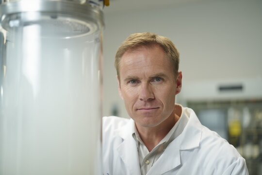 Male research scientist in a white lab coat stands in a laboratory with glassware.