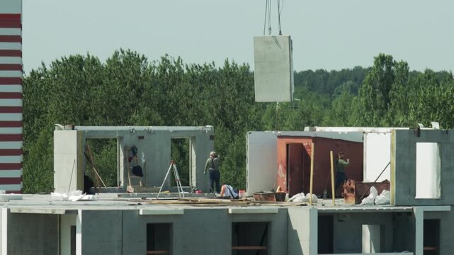 Crane Lifts Concrete Panel Over Construction Site. Workers On Partially Built Upper Floor.