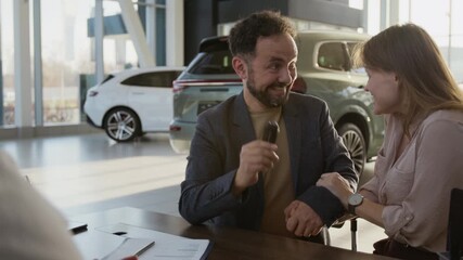 Medium shot of smiling adult couple signing contract and embracing happily while buying new car in dealership showroom with salesman giving keys