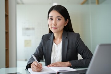 Professional businesswoman sits at a desk, writing notes in a modern office.