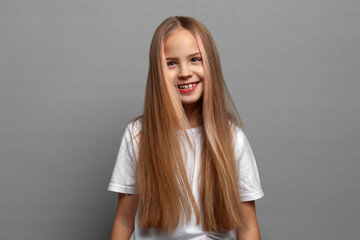 Close up portrait of beautiful happy smiling blonde girl with long hair in white clothing. Cute young child posing on gray studio background with natural joyful expression