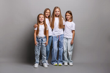 Full length portrait happy children, preteen girls with long hair standing together in studio, smiling and hugging, wearing white t-shirts and jeans, friendship, childhood, unity, joy and kids fashion