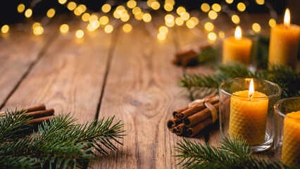 Warm and cozy Christmas ornaments on a rustic wooden table top decorated with pine branches, cinnamon sticks, and lit candles, with blurred golden fairy lights in the background.