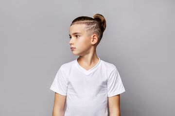 Young boy with trendy shaved hairstyle and topknot looking aside in studio, wearing plain white t-shirt, modern stylish portrait isolated on neutral background