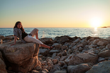 A girl poses and relaxes by the sea, sitting on rocks near the slope of a high cliff, panoramic view of the seashore and mountains at sunset