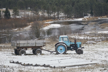 An old tractor with a cart in winter in a field with snow and logs near the river