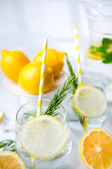 Glasses of lemon water with rosemary sprigs yellow striped straws and fresh lemon halves arranged with mint and ice