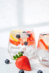 Fresh summer drink with lemon slices strawberries and blueberries served in clear glass with ice on light background