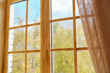 Open wooden window frame overlooking a forest with trees and blue sky and a sheer curtain blowing in