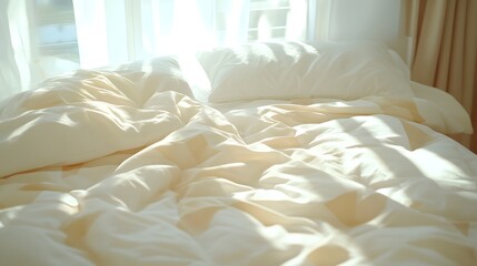Unmade bed covered in light colored bedding receives bright morning sunlight through a window.